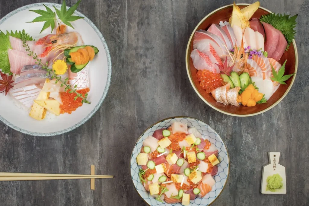 A top-down view shows three colorful Japanese rice bowls arranged on a dark, textured surface along with chopsticks and a small paddle with wasabi. Two bowls are artfully topped with sliced raw fish, uni, shrimp, tamago, and salmon roe, while the third features a diced scattered chirashi layout.