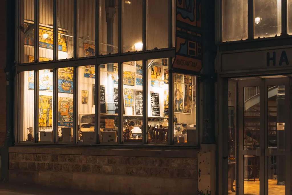 The image captures a brightly lit cafe or museum interior at night, viewed from the outside through large glass windows. The walls inside are adorned with colorful, naive-style artwork and menu boards, while a single person is visible working behind the counter.