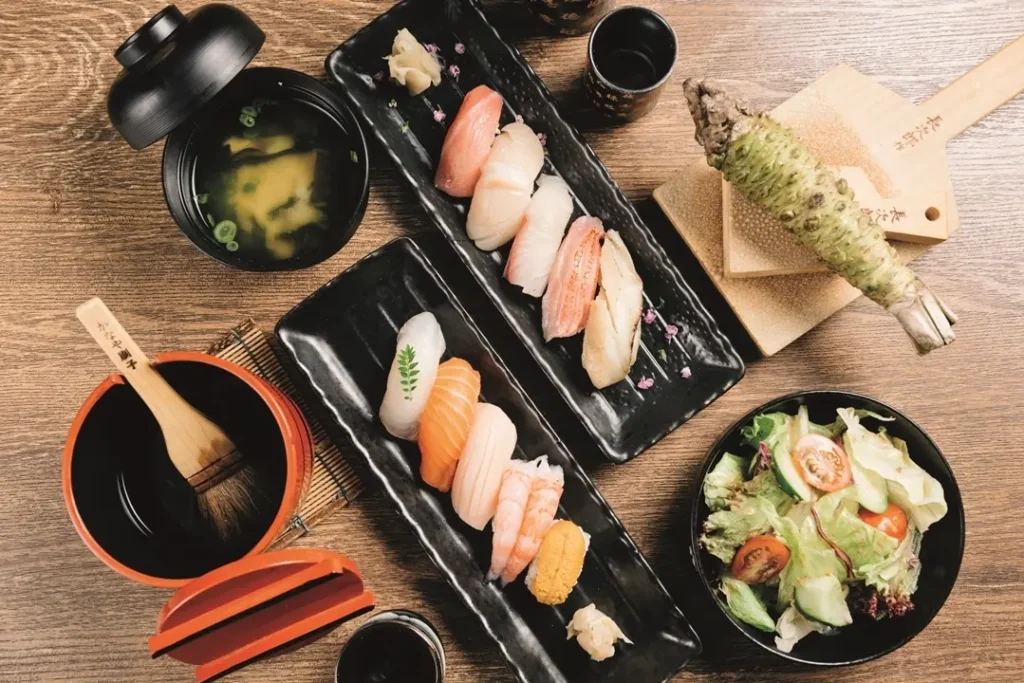 An overhead view displays two rectangular black platters arranged with various types of fresh nigiri sushi on a wooden tabletop. Surrounding the sushi are side dishes, including a bowl of green salad with tomatoes, a lidded bowl of miso soup, soy sauce, and a whole fresh wasabi root with a wooden grater.