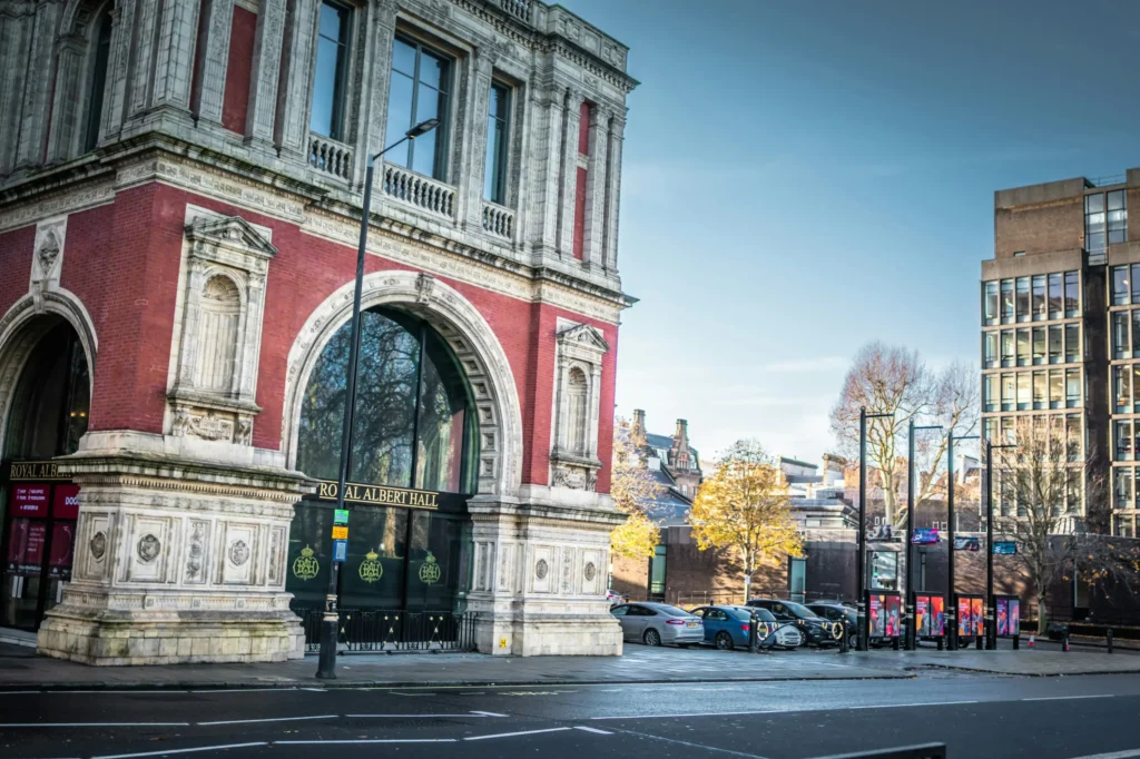 The image captures a side view of the historic, red brick and ornate white stone facade of the Royal Albert Hall in London under a clear blue sky. In the background, parked cars line the street adjacent to modern buildings and trees with autumn foliage.