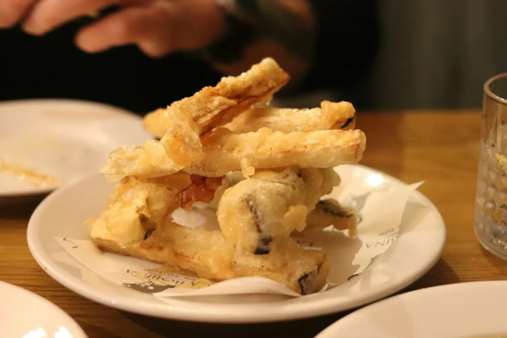 A small plate of crispy, golden-brown tempura-battered eggplant fries is stacked neatly on a white dish lined with paper. The dish sits on a wooden table in a dimly lit restaurant setting, with hands partially visible in the background.