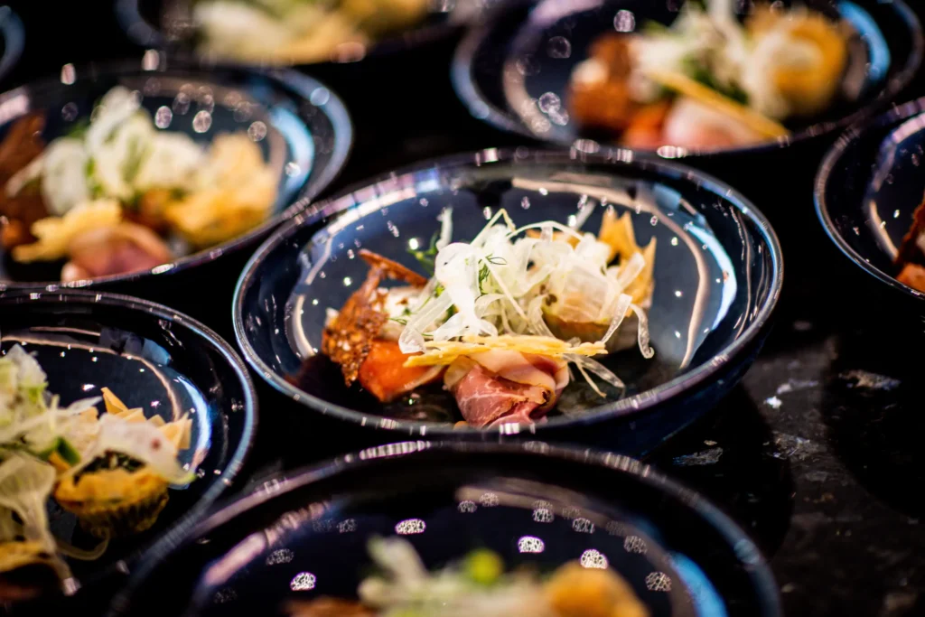 A dark stone bowl filled with white rice is artfully topped with shredded seaweed, yellow pickled radish, bright orange fish roe, and microgreens. The dish sits on a wooden restaurant table with a bowl of soup visible in the background.