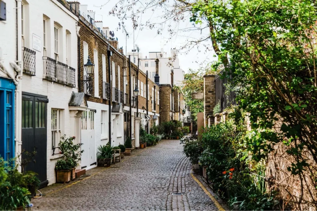 A narrow, charming cobblestone mews street is lined with historic multi-story terraced houses featuring white and brown brick facades. Lush green plants, potted flowers, and overhanging trees frame the walkway, adding a vibrant, picturesque touch to the quiet lane.