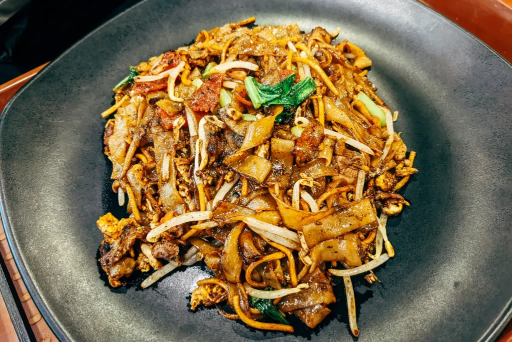 A high-angle shot shows a large plate of Char Kway Teow, a stir-fried noodle dish, served on a dark, textured round plate. The dish features a mix of flat rice noodles, yellow noodles, bean sprouts, scrambled egg, and green vegetables coated in a dark, glossy savory sauce.
