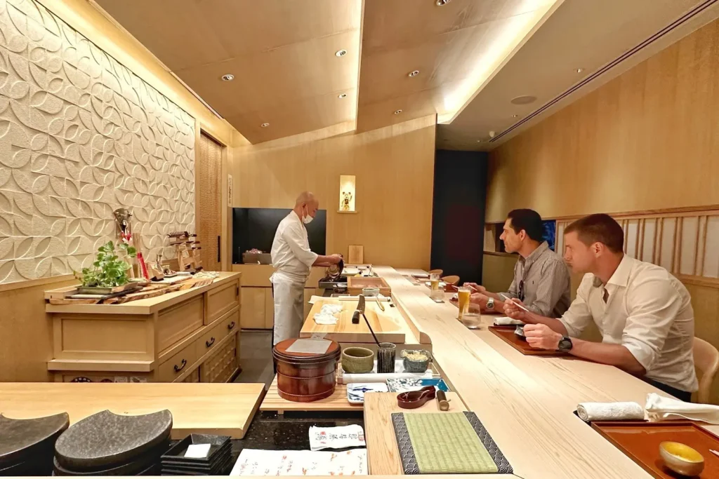 A Japanese sushi chef in a chef's uniform works behind a long, light-wood dining counter in a warmly lit, modern minimalist restaurant. Two male diners sit across from him at the bar, watching attentively as he prepares food.