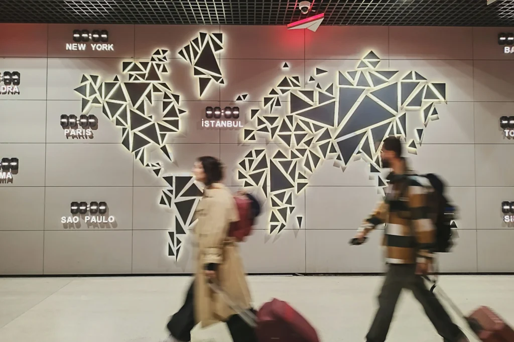 A large, backlit geometric world map made of dark triangles is mounted on a gray tiled wall, surrounded by digital clocks displaying major city names like New York and Istanbul. In the foreground, two blurred travelers walk past the display while pulling their luggage, capturing a sense of movement.