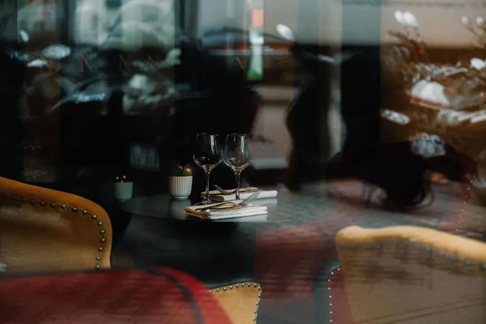 The image shows a dimly lit restaurant interior captured through a glass window, creating a layered effect with reflections of the outside environment. On a small round table, two wine glasses stand alongside neatly folded white napkins with silverware, flanked by plush yellow and red armchairs.