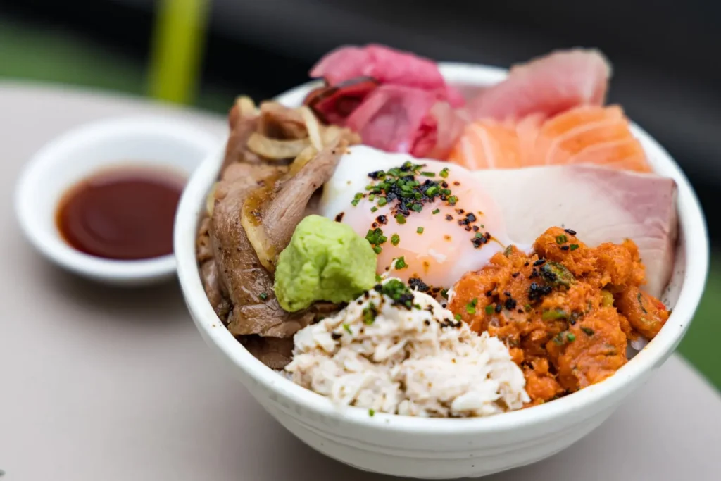 A detailed Japanese donburi rice bowl filled with sliced beef, fresh sashimi, spicy tuna, crab meat, pickled ginger, and a soft-poached egg topped with herbs. A dollop of wasabi rests on the side, accompanied by a small dish of soy sauce in the background.