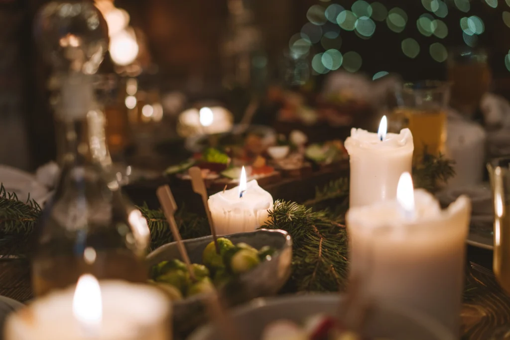 The image depicts a warm, dimly lit festive dinner table decorated with thick, burning white candles and green pine sprigs. Various bowls of holiday appetizers and glassware are scattered across the table, set against a soft background of blurred green bokeh lights.