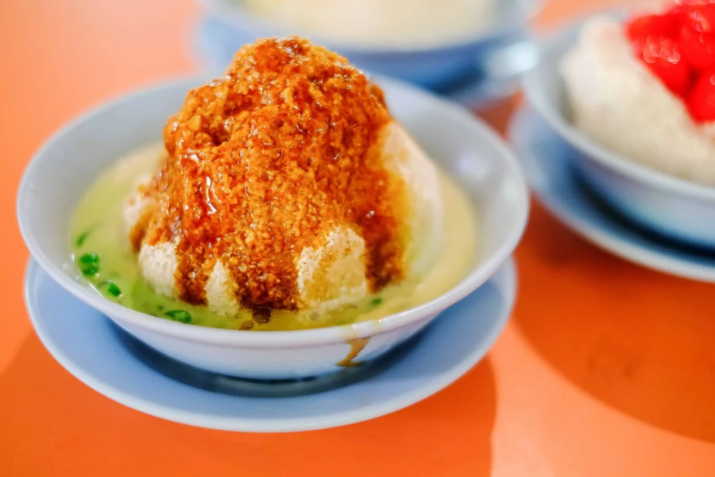 A close-up shot of a bowl of Cendol, a traditional Southeast Asian dessert featuring a mound of shaved ice drenched in dark brown palm sugar syrup. The ice sits in a light green coconut milk broth with small green rice flour jellies, served on an orange tabletop with other bowls blurred in the background.