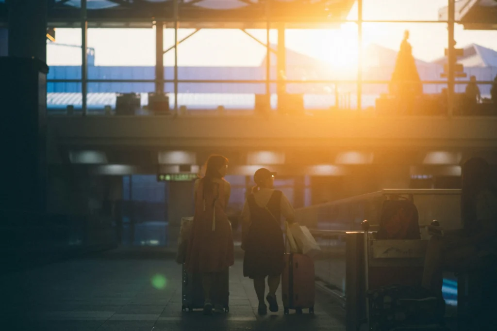 Two travelers with rolling luggage walk away from the camera through a dimly lit airport terminal or transit hub. Intense, warm golden sunlight streams through the large glass windows ahead, creating a strong contrast and lighting up the upper level of the building.