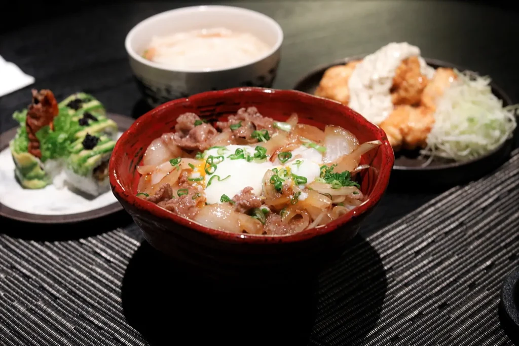 A Japanese beef rice bowl topped with a soft-boiled egg and green onions sits in a red bowl on a dark textured placemat. Surrounding the main dish are side plates containing sushi rolls topped with avocado, a dish with creamy sauce, and a small bowl of soup.