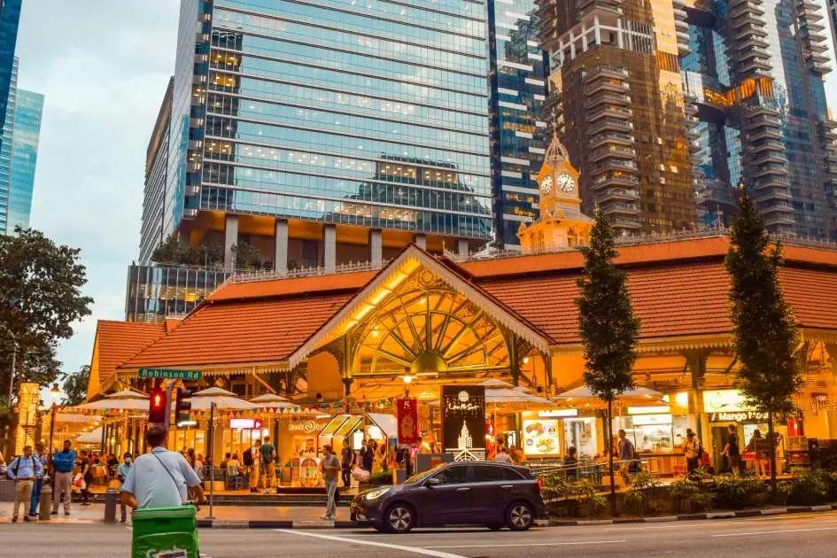 A bustling street view features the historic Lau Pa Sat hawker center in Singapore, illuminated by warm lights with its distinctive tiled roof, clock tower, and outdoor dining stalls. In the background, towering modern glass and metal skyscrapers rise sharply against the evening sky, creating a striking architectural contrast.