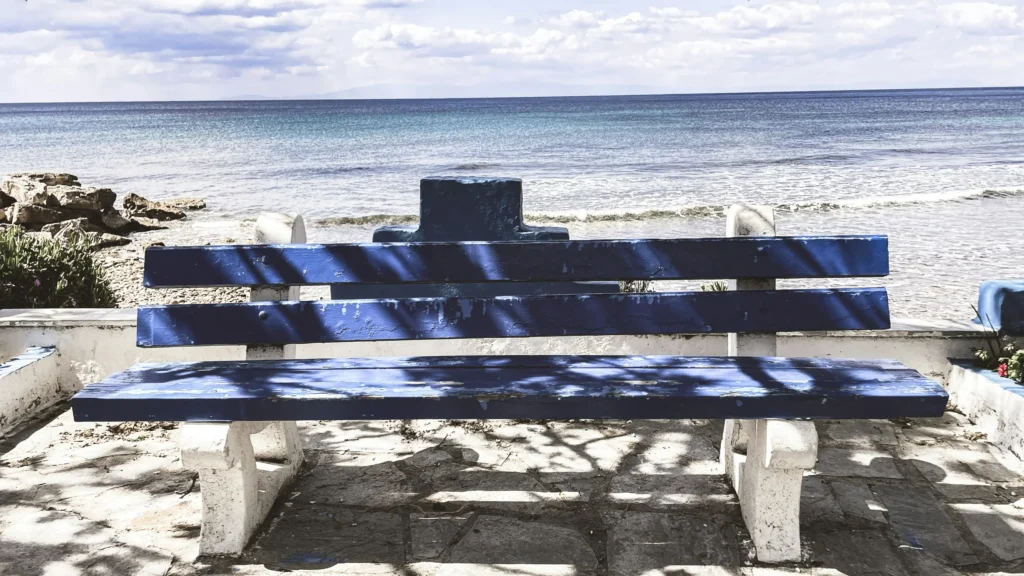 A painted blue wooden bench with white concrete legs sits on a stone patio overlooking a calm ocean view. Soft dappled shadows from tree branches above rest on the bench and paved ground under a bright, partly cloudy sky.