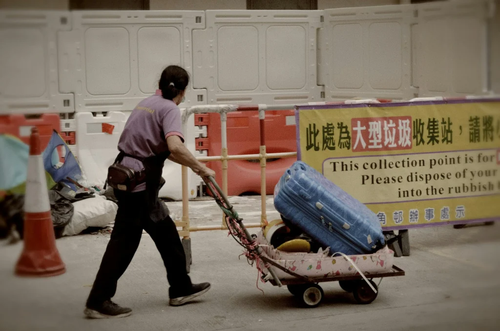 In the image hayley-kee-hqXkXPBJFKo-unsplash.jpg, a person wearing a purple shirt and dark pants pulls a small cart loaded with a large blue suitcase. They are walking past a setup of white and red construction barriers next to a yellow sign indicating a collection point.