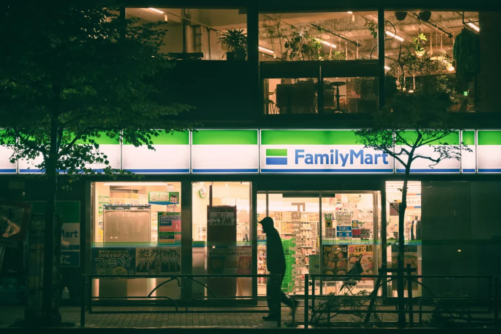 A brightly lit FamilyMart convenience store illuminates a dark street at night, its signature green and white logo glowing above the glass windows. A silhouetted figure wearing a hoodie walks past the warm, inviting light of the entrance.