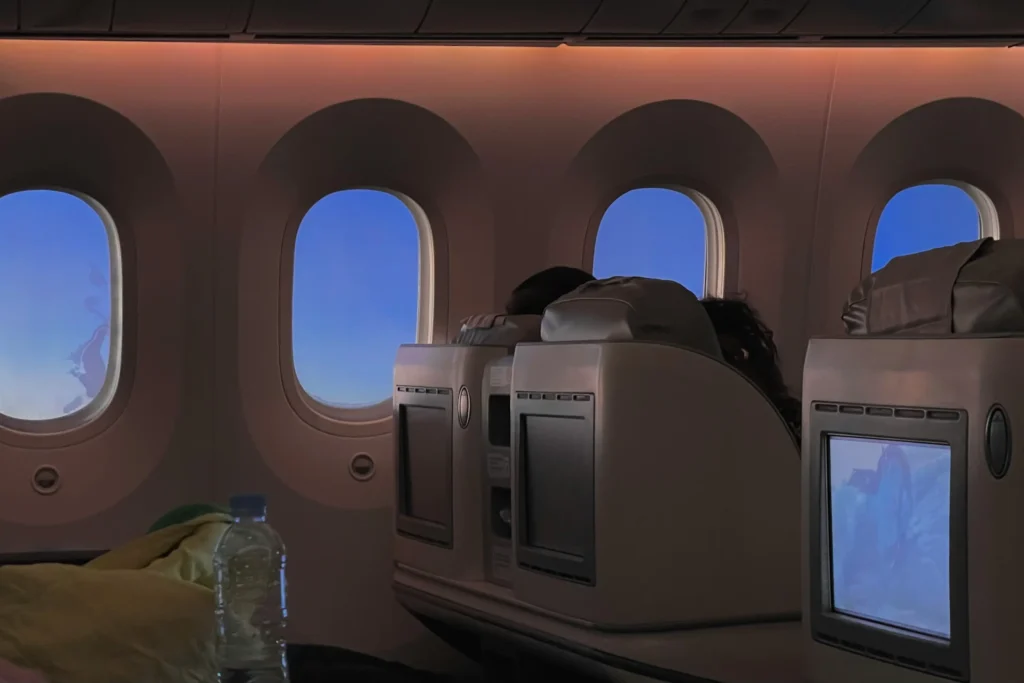 An interior view of an airplane cabin shows passengers seated in premium or business class seats with personal entertainment screens. A row of large, oval windows looks out onto a clear, bright blue sky during a daytime flight.