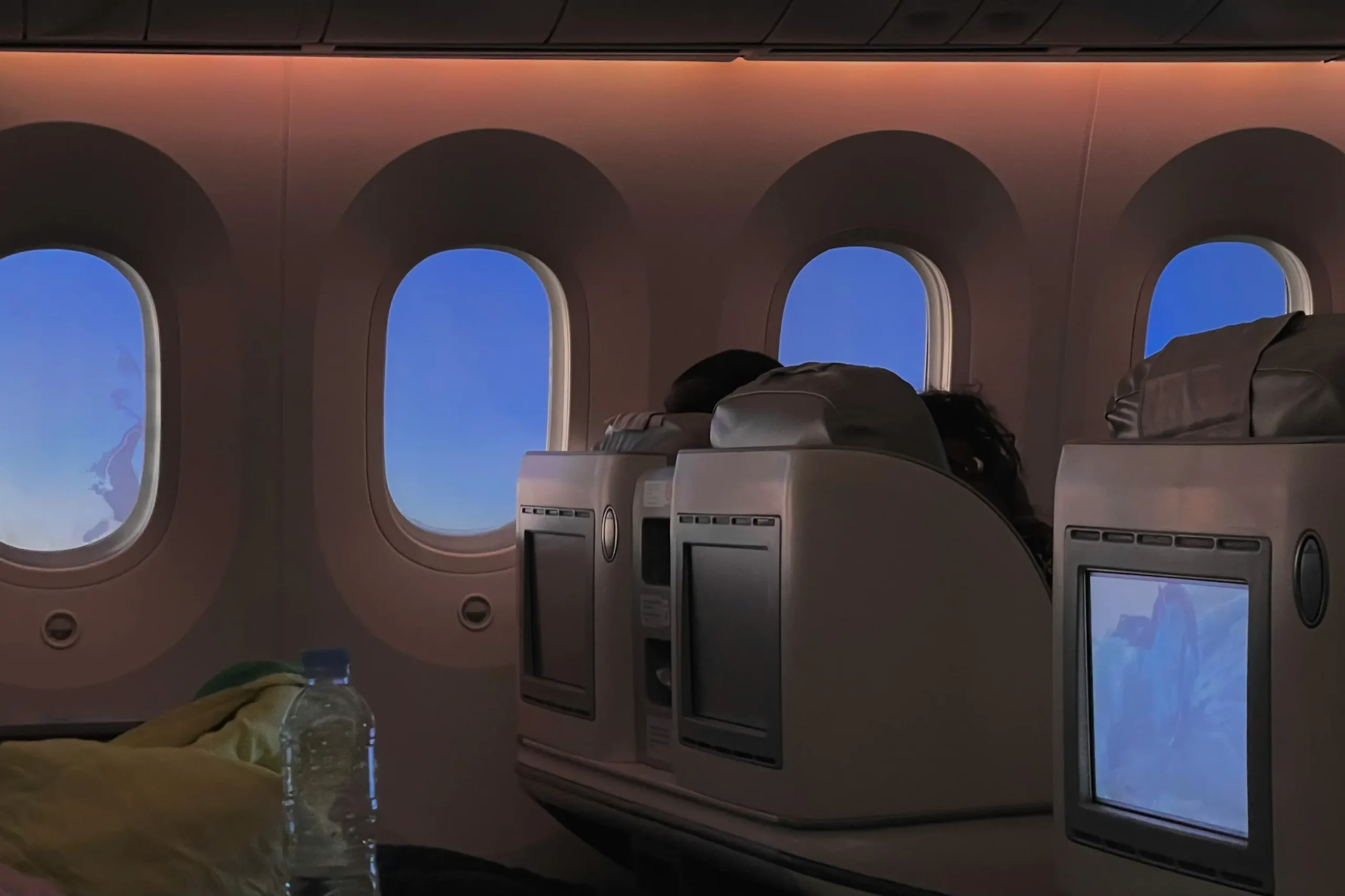 An interior view of an airplane cabin shows passengers seated in premium or business class seats with personal entertainment screens. A row of large, oval windows looks out onto a clear, bright blue sky during a daytime flight.