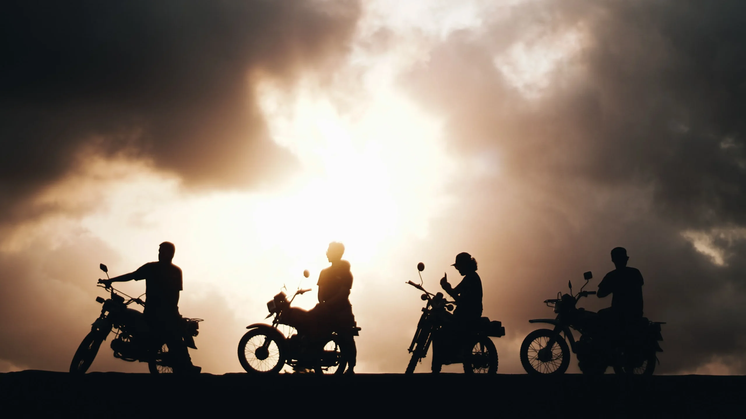 The image shows a silhouette of four motorcyclists lined up against a dramatic, cloudy sky during sunset or sunrise. The bright sun breaks through heavy storm clouds, casting a warm glow and sharply contrasting with the dark figures on their bikes.