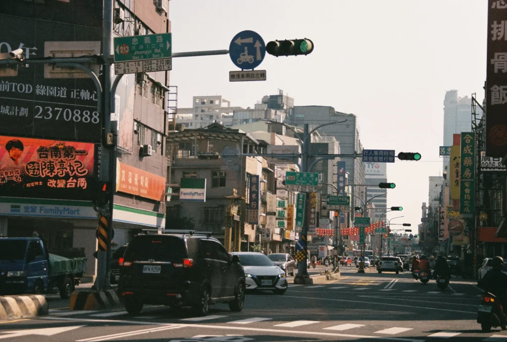 A bustling city intersection in Taiwan features cars and motorbikes navigating a street lined with multi-story buildings, storefronts like FamilyMart, and vibrant billboards. Overhead, green traffic lights and various blue and green road signs, including one pointing to Jhongyi Road, hang above the multi-lane roadway.