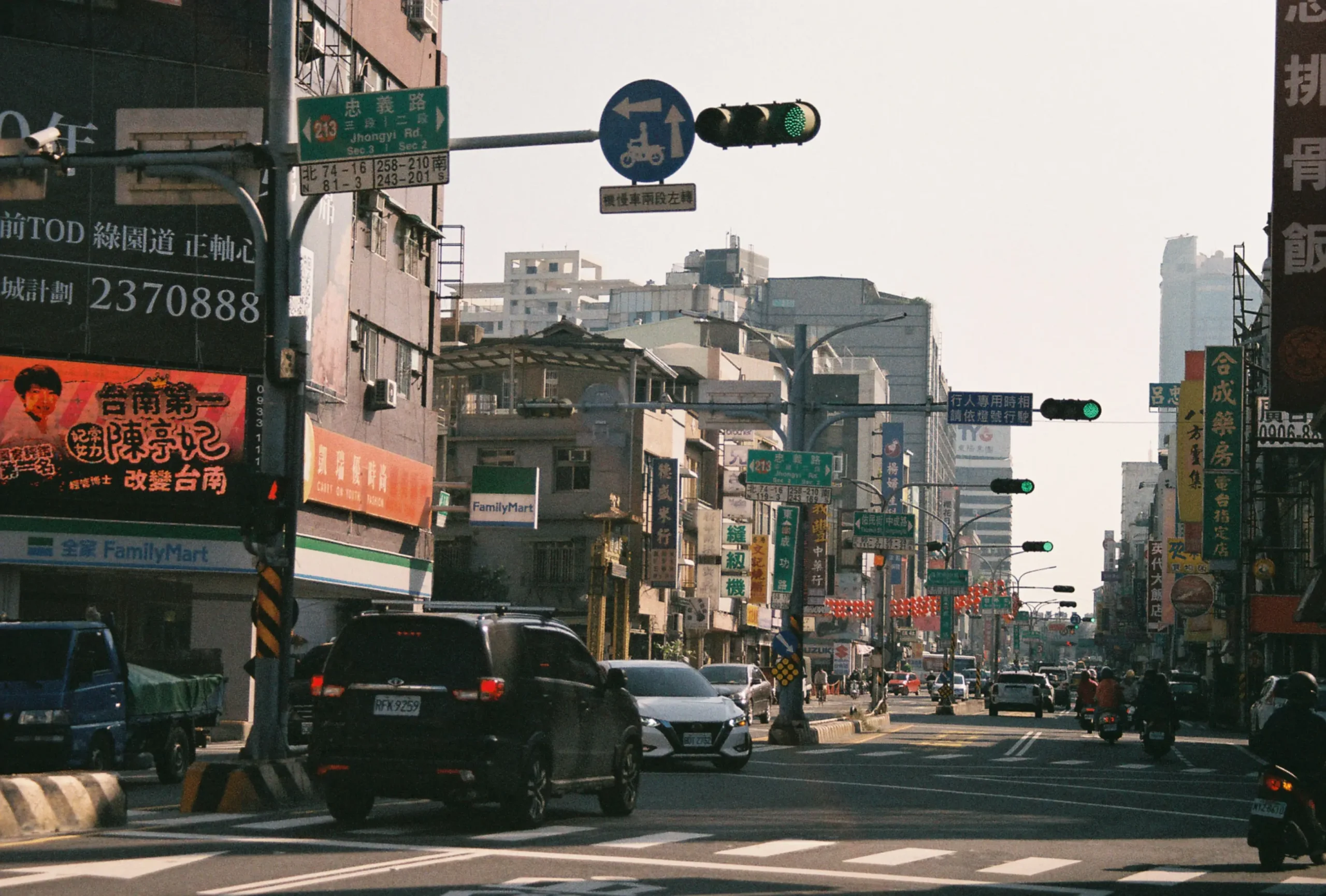 A bustling city intersection in Taiwan features cars and motorbikes navigating a street lined with multi-story buildings, storefronts like FamilyMart, and vibrant billboards. Overhead, green traffic lights and various blue and green road signs, including one pointing to Jhongyi Road, hang above the multi-lane roadway.