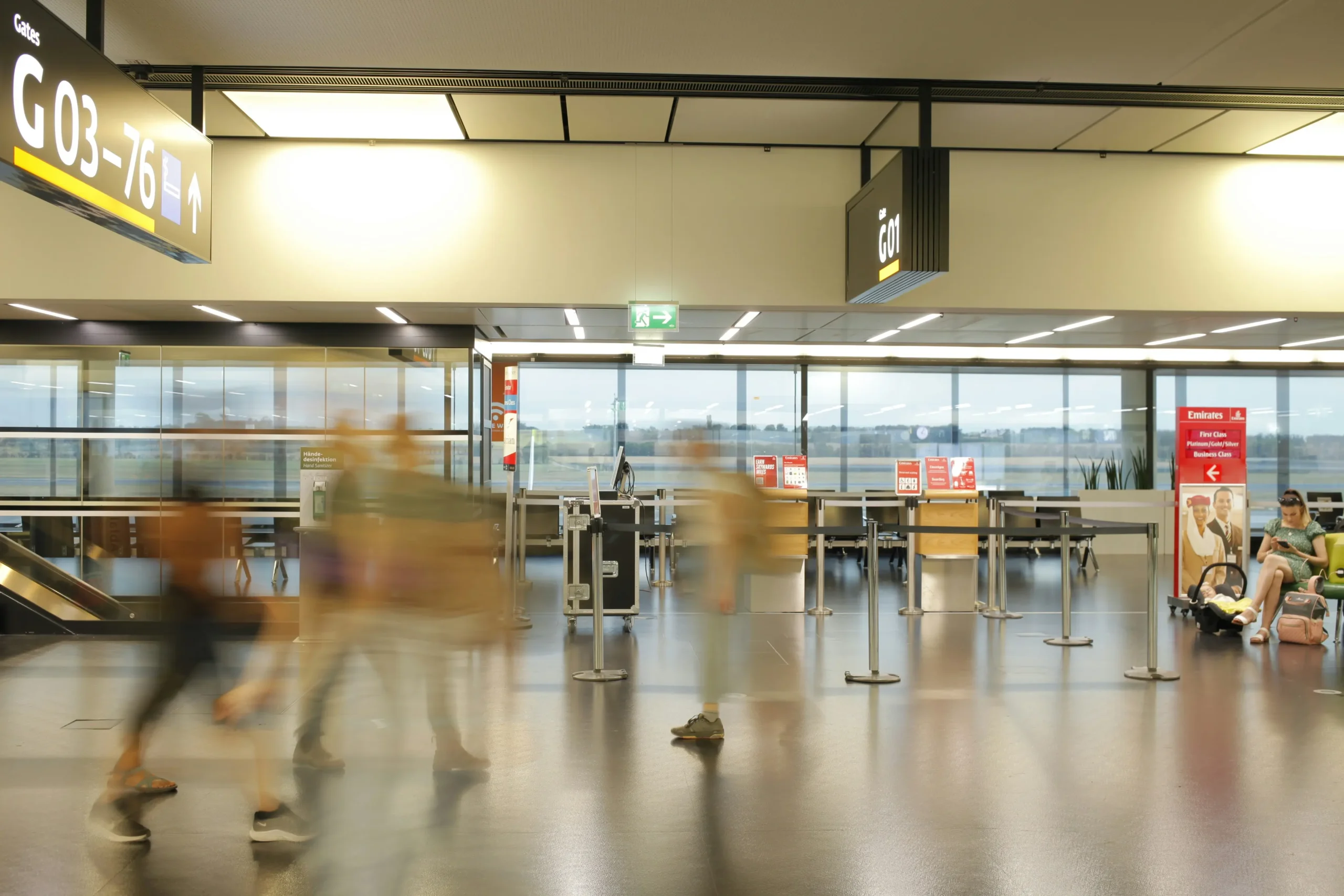 A wide shot of a modern, brightly lit airport terminal with blurred figures of people walking through the concourse, capturing a sense of motion. Large signs indicate gate numbers "G03-76" and "G01" above empty check-in counters and stanchions lined up against a wall of windows.