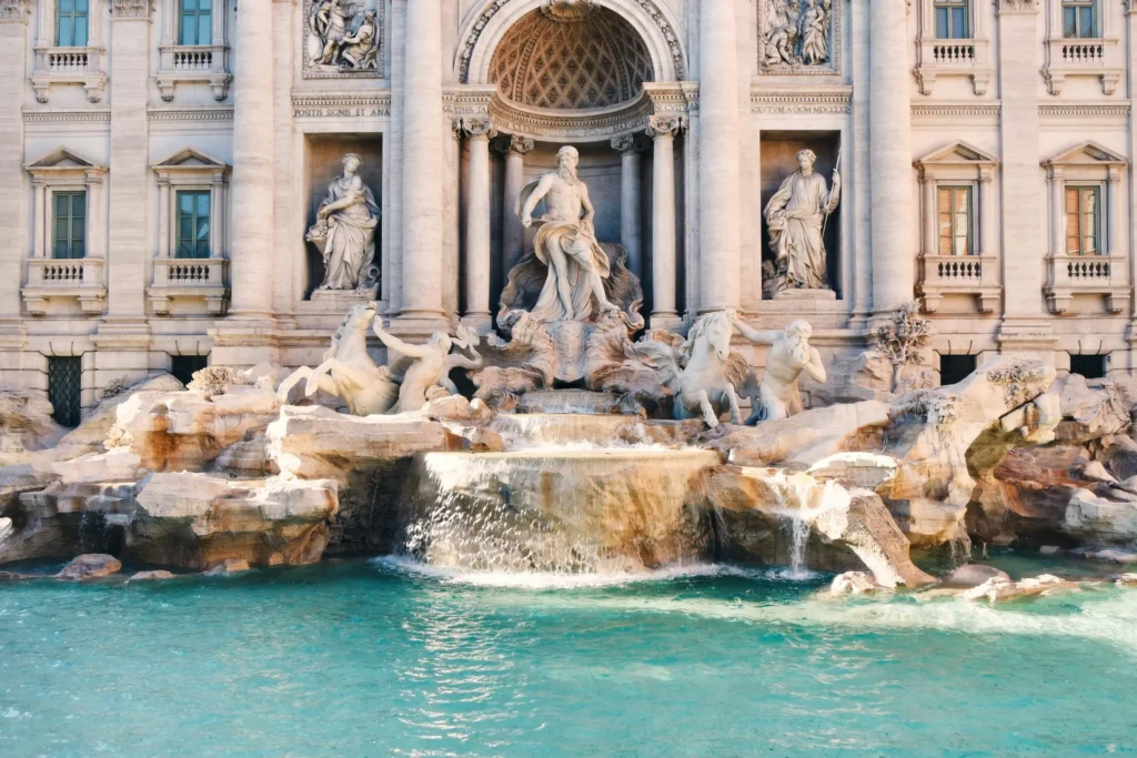 The majestic Trevi Fountain in Rome features intricate marble sculptures of Oceanus and mythological figures set against a grand baroque fau00e7ade. Sunlight illuminates the crystal-clear turquoise water cascading over the natural stone rockwork into the basin below.