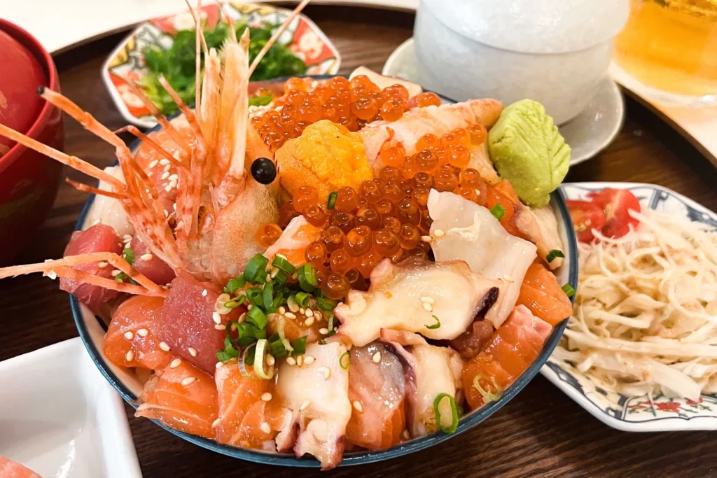 A close-up view of a vibrant Japanese Omote rice bowl overflowing with fresh seafood, including whole shrimp, salmon, tuna, octopus, uni, and shiny salmon roe. The bowl is garnished with scallions, sesame seeds, and a dollop of wasabi, set alongside side dishes on a wooden table.