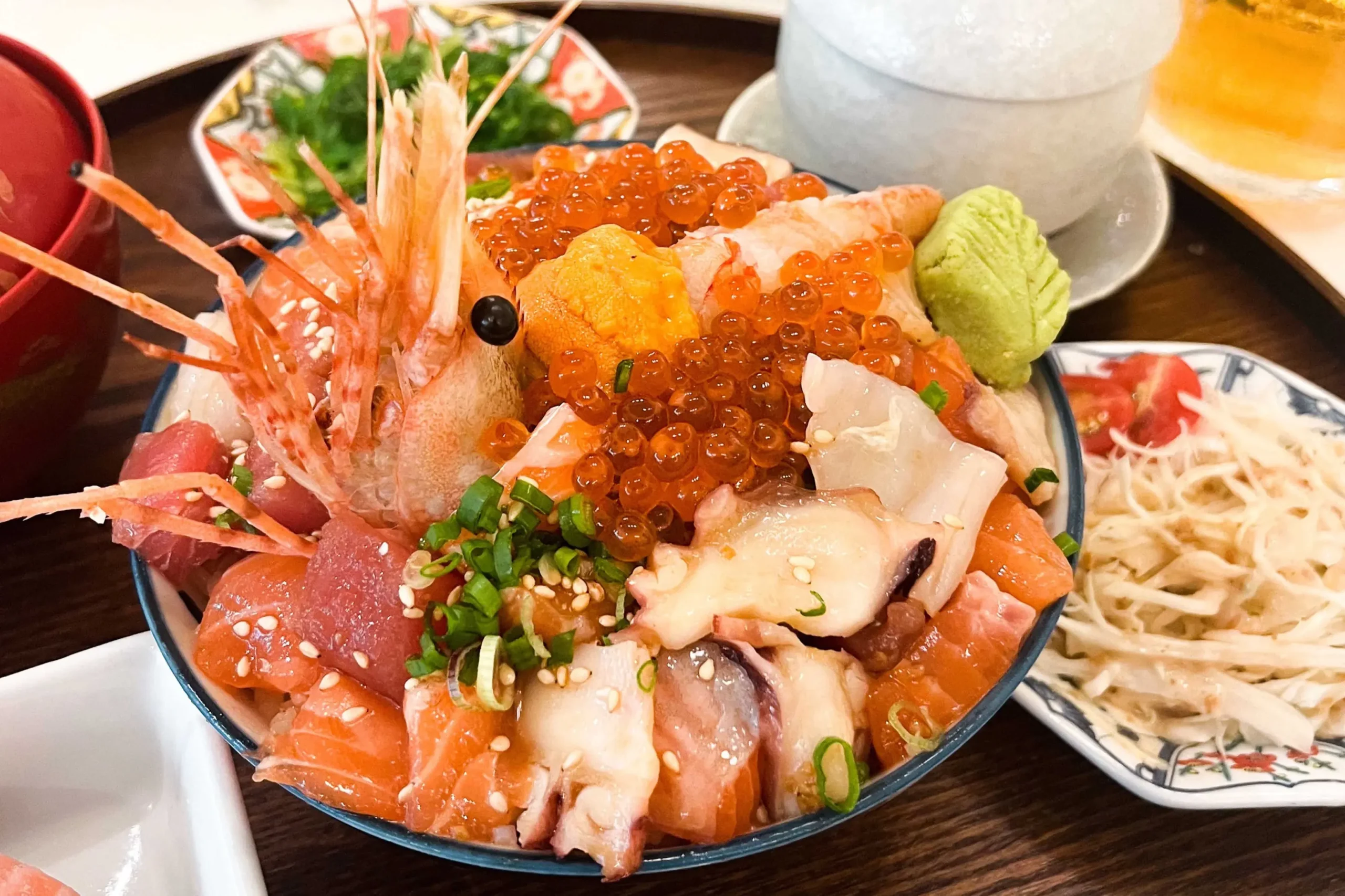A close-up view of a vibrant Japanese Omote rice bowl overflowing with fresh seafood, including whole shrimp, salmon, tuna, octopus, uni, and shiny salmon roe. The bowl is garnished with scallions, sesame seeds, and a dollop of wasabi, set alongside side dishes on a wooden table.