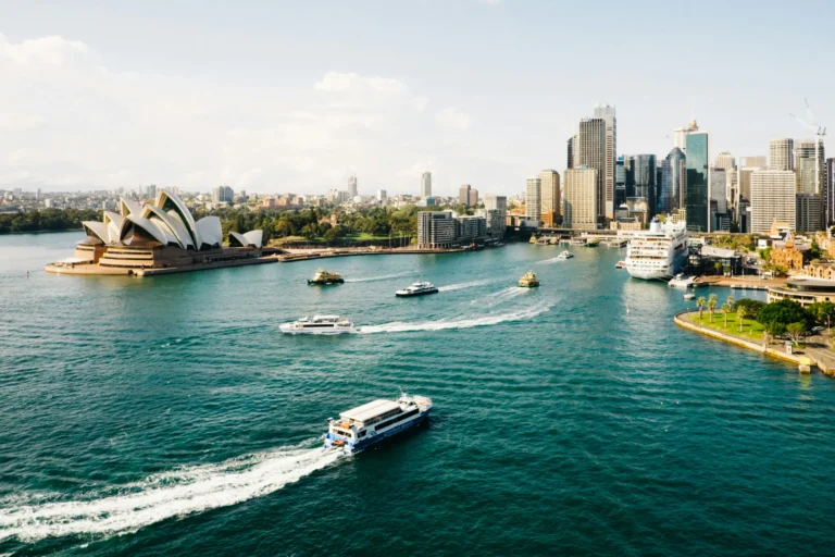 An aerial view of Sydney Harbour showing the iconic Sydney Opera House on the left shoreline and city skyscrapers on the right. Ferries and boats navigate through the bright blue water under a clear sky.