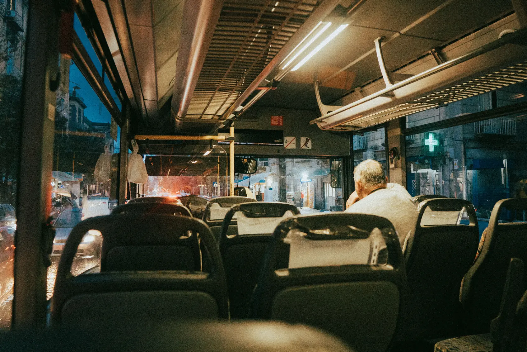 The interior of a dimly lit bus captured with film grain shows rows of empty seats leading toward the front as it travels along a city street at night. A lone passenger with gray hair sits near the window on the right, looking out at glowing streetlights and city buildings.