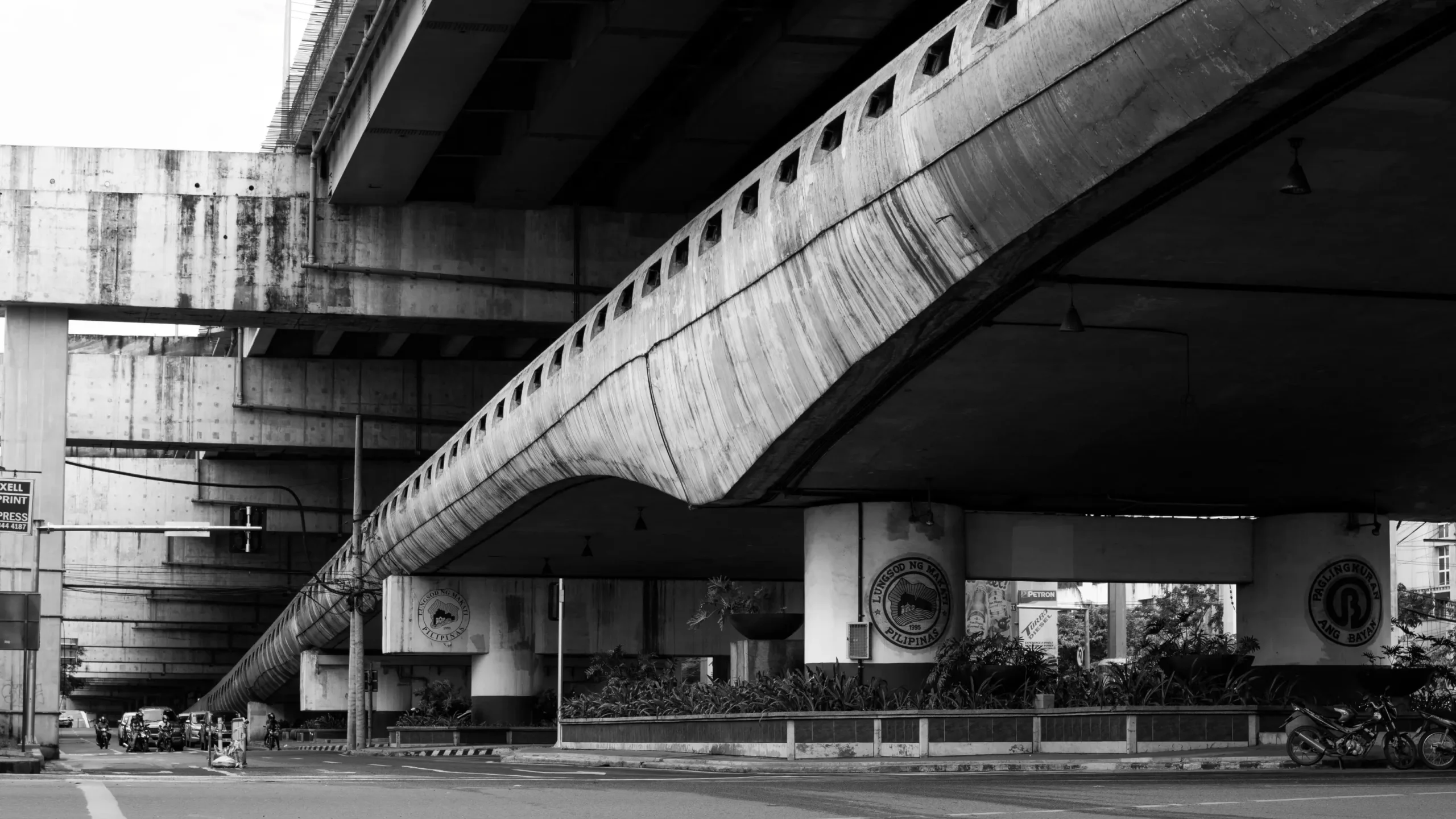 A black-and-white view beneath a massive elevated concrete highway and overpass structure with supportive pillars. Below the bridge, a street with light traffic and landscaped planter beds is visible.