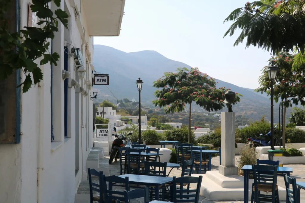 A sunny outdoor patio features blue wooden tables and chairs set against whitewashed Mediterranean-style buildings. In the background, lush green trees and a prominent bust statue stand before scenic mountain hills.