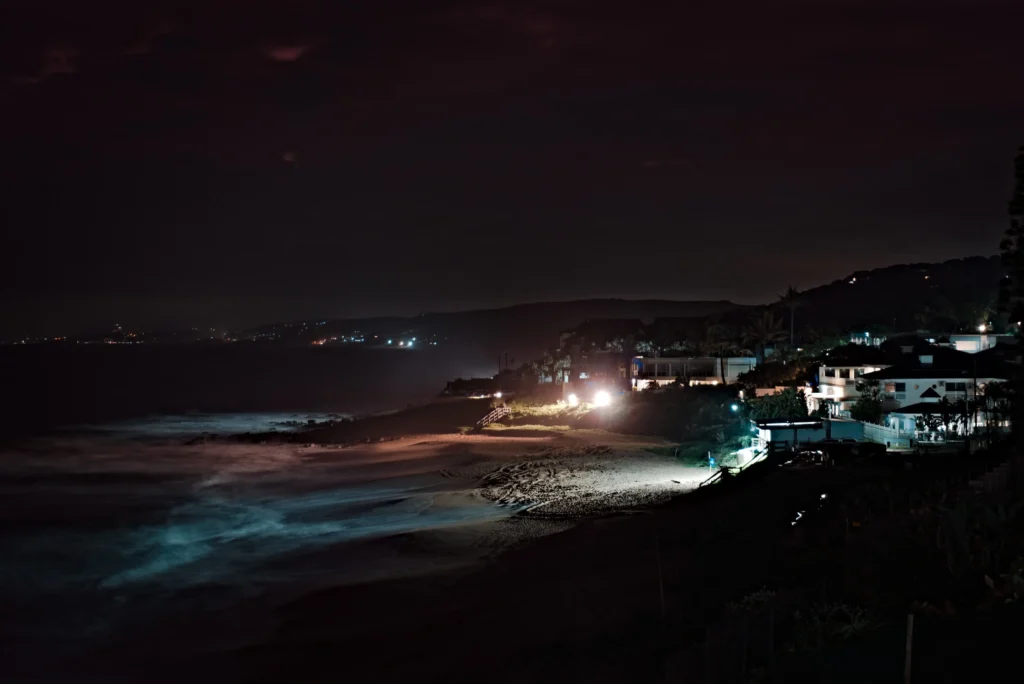 A nighttime long-exposure shot captures gentle ocean waves glowing under dim artificial light along a sandy shoreline. Coastal houses and streetlights illuminate the dark hillside and beach in the background.