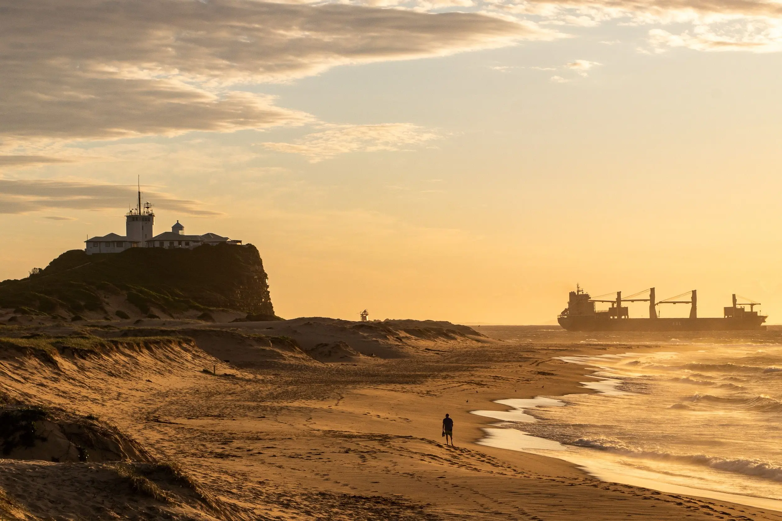 A solitary person walks along a wide sandy beach bathed in warm golden sunlight during early morning or late afternoon. In the background, a lighthouse stands atop a grassy hill overlooking the ocean, while a large cargo ship sails on the horizon.