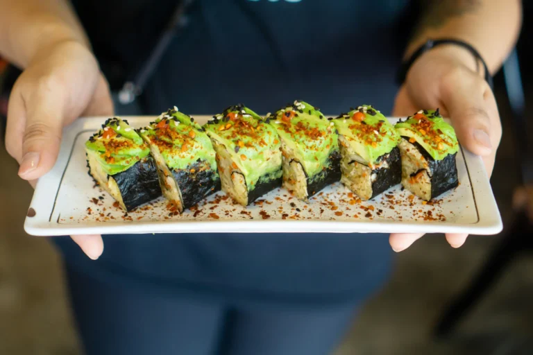 A person holds a long white rectangular plate filled with a row of six sliced sushi rolls wrapped in dark nori. Each roll is topped with fresh sliced avocado, black and white sesame seeds, red chili flakes, and small dots of sauce.