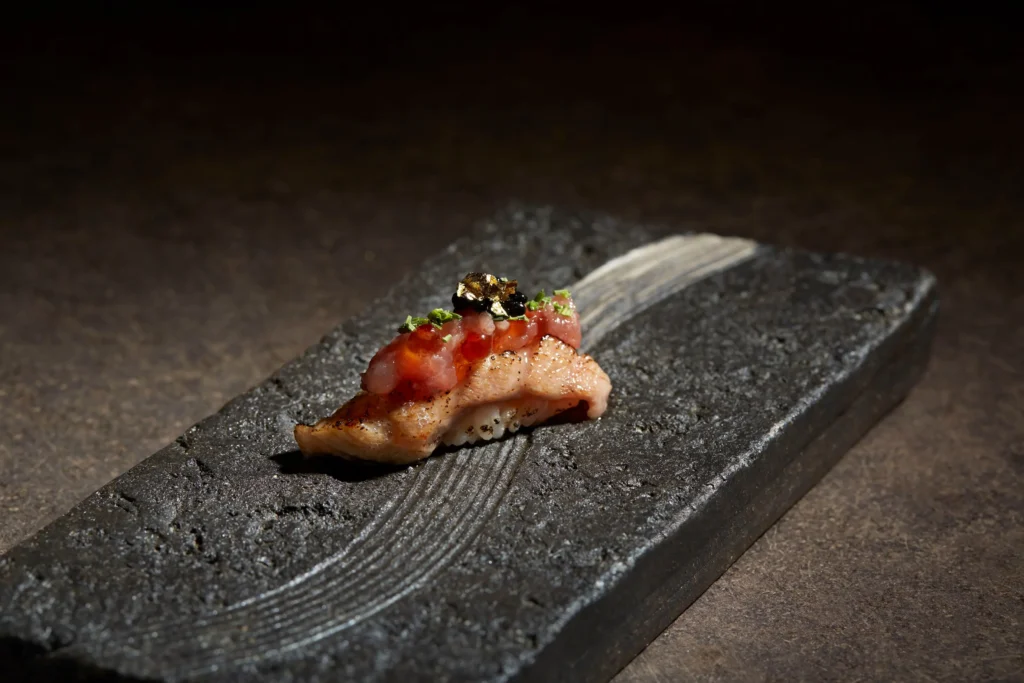 A piece of gourmet seared nigiri sushi rests on a dark, textured stone platter under dramatic lighting. It is topped with minced raw fish, glistening salmon roe, black caviar, herbs, and a delicate speck of edible gold leaf.