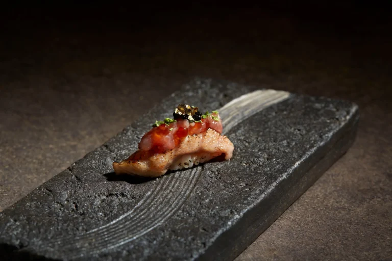 A piece of gourmet seared nigiri sushi rests on a dark, textured stone platter under dramatic lighting. It is topped with minced raw fish, glistening salmon roe, black caviar, herbs, and a delicate speck of edible gold leaf.