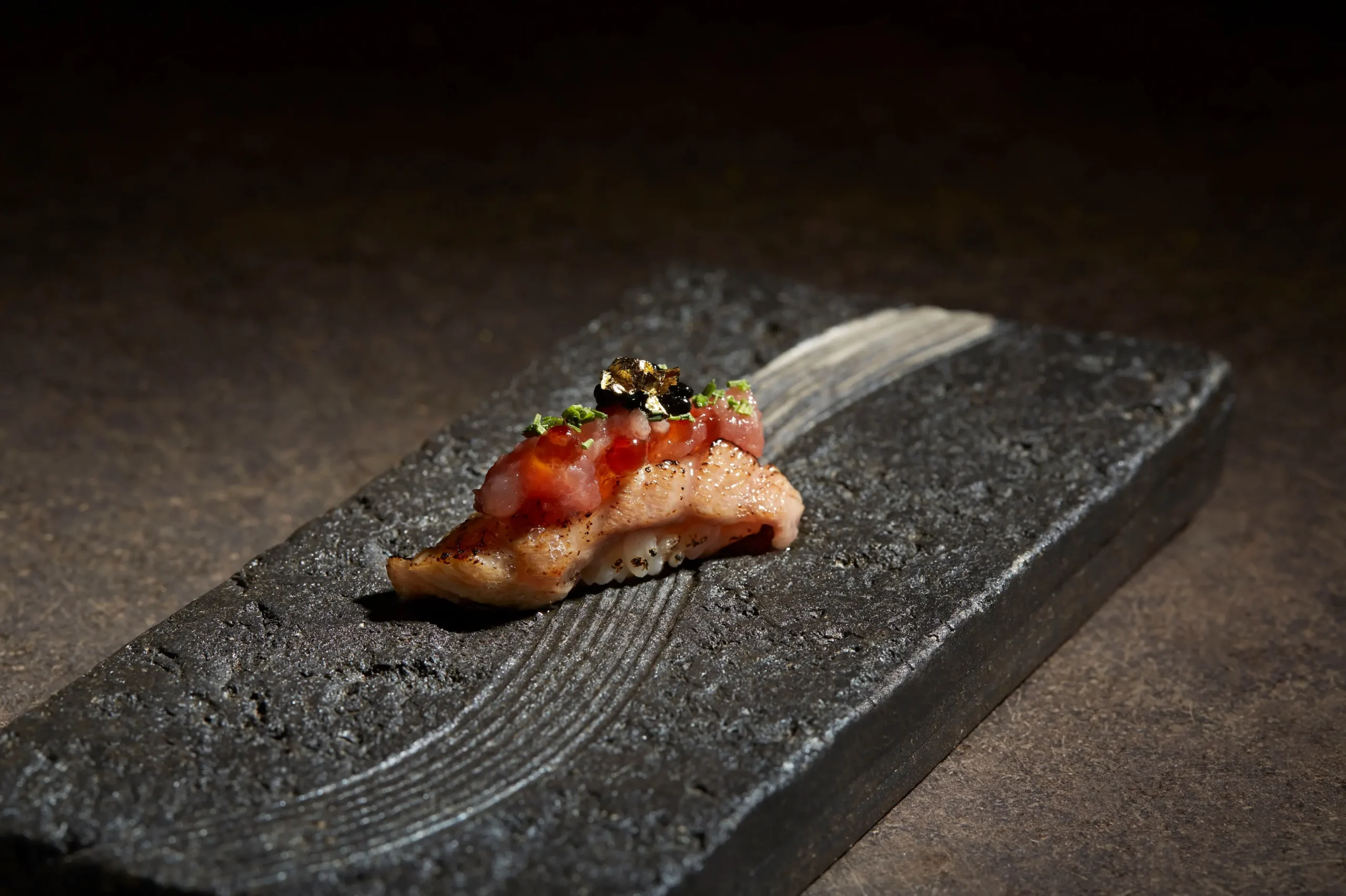 A piece of gourmet seared nigiri sushi rests on a dark, textured stone platter under dramatic lighting. It is topped with minced raw fish, glistening salmon roe, black caviar, herbs, and a delicate speck of edible gold leaf.
