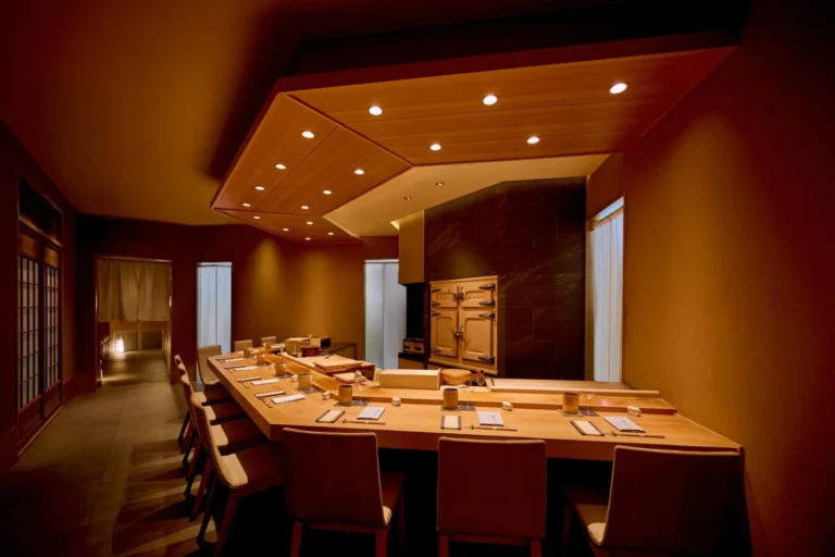 An elegant, dimly lit Japanese dining room features a long, smooth wooden sushi counter set with matching chairs. Warm overhead lighting casts a cozy glow over the neatly arranged place settings along the bar.