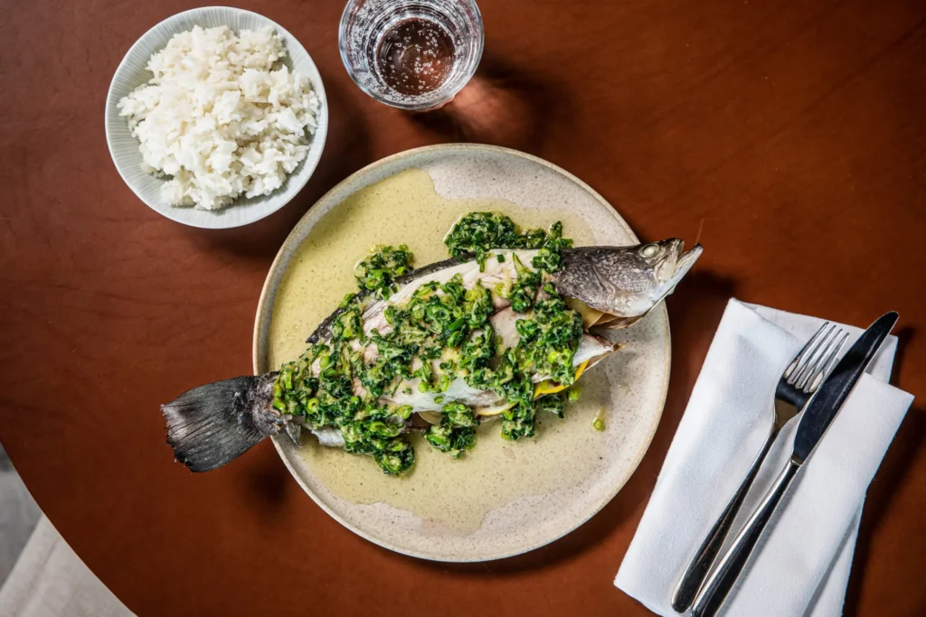 A whole cooked fish topped with bright green herbs sits on a plate on a wooden table. Beside it, there is a bowl of white rice, a glass of water, and cutlery neatly wrapped in a cloth napkin.