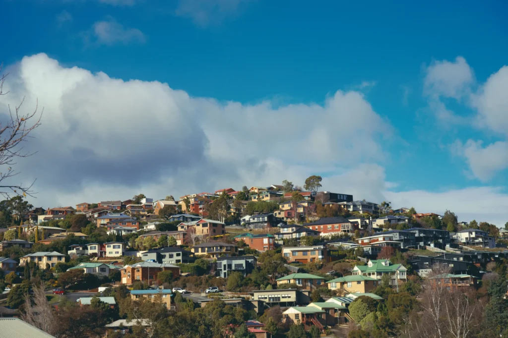 A hillside suburban neighborhood filled with multi-story houses nestled among green trees and vegetation. Above, large white fluffy clouds drift across a bright blue sky.