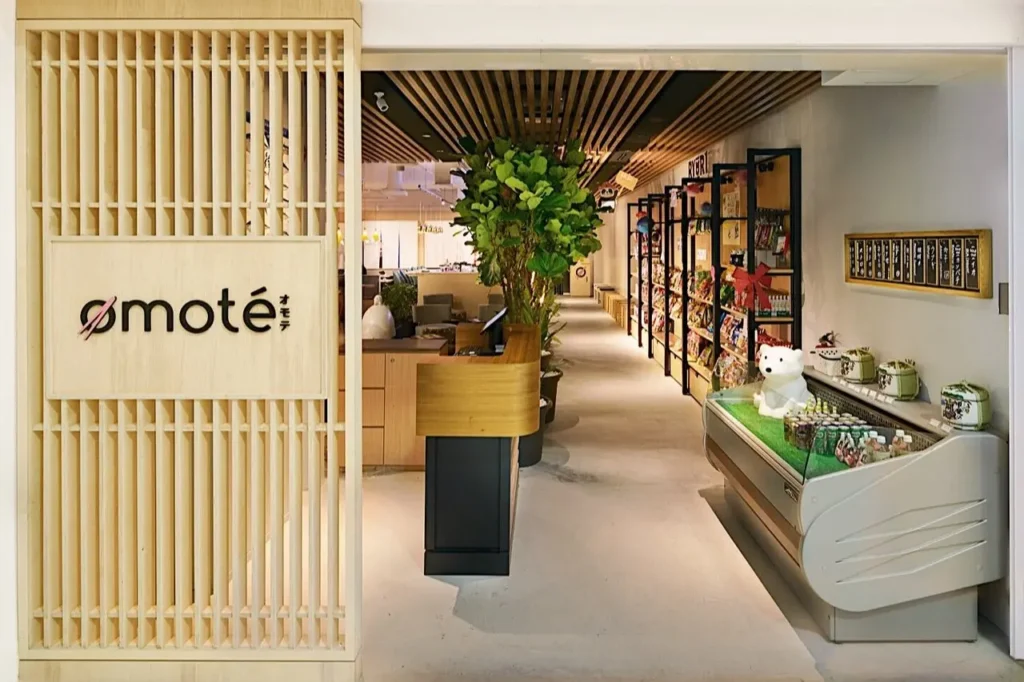 The modern, light wood entrance to a restaurant called Omote features a vertical slatted divider displaying the logo on the left. Inside, a hallway extends past a wooden reception counter, lush potted plants, merchandise shelves, and a refrigerated display case filled with drinks.
