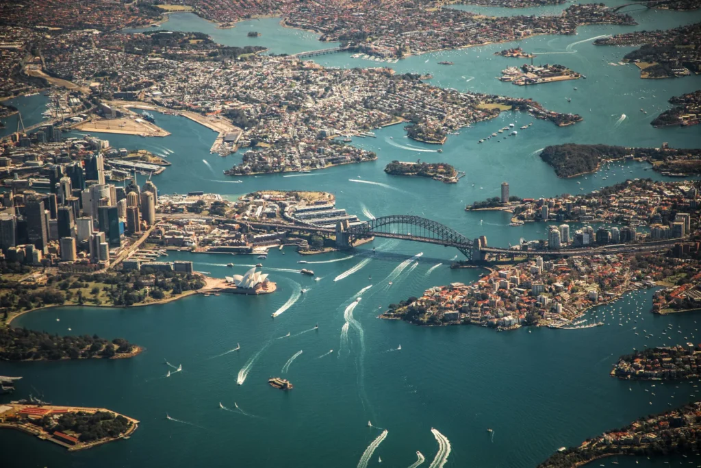 An aerial view captures the sprawling city of Sydney and its iconic harbor, featuring the Sydney Harbour Bridge and the Sydney Opera House. Numerous boats leave white wakes as they navigate the deep blue waters surrounding the urban coastline.