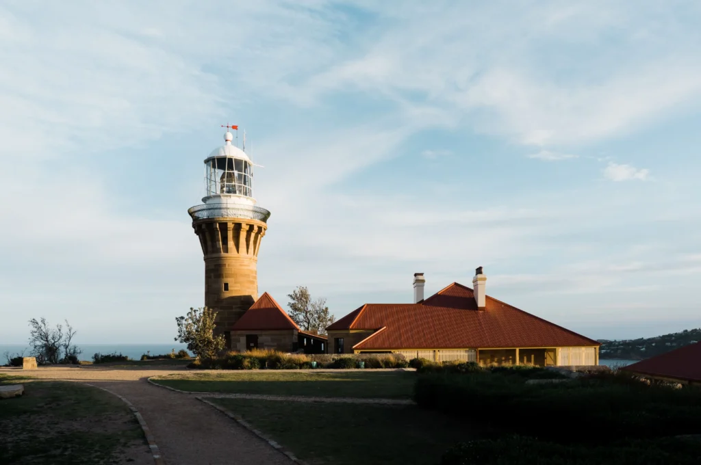 A historic stone lighthouse stands beside a building with a bright red roof on a grassy coastal headland. A dirt path winds past the lighthouse grounds, overlooking the ocean under a clear, pale blue sky.