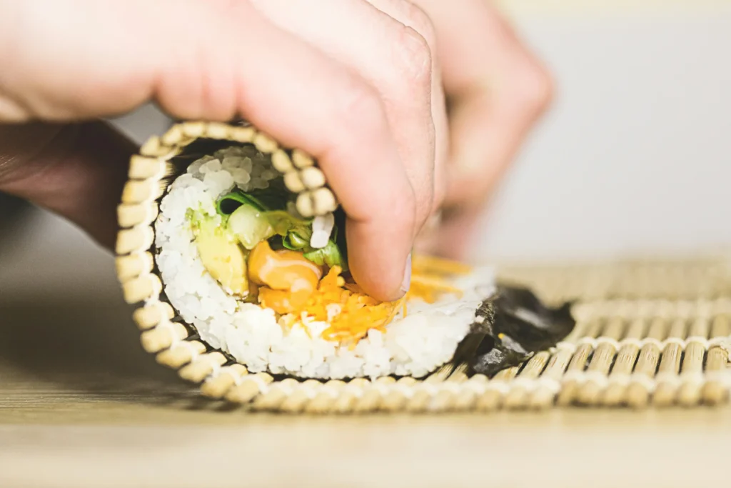 A pair of hands carefully rolls a homemade sushi roll using a traditional bamboo rolling mat. The cross-section reveals fillings of sushi rice, fresh salmon, avocado, greens, and shredded carrots wrapped in nori seaweed.