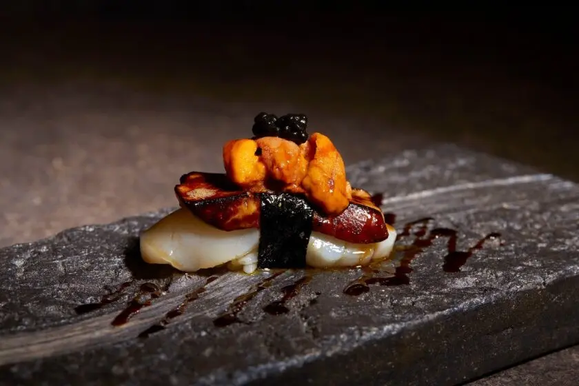 A piece of gourmet nigiri sushi is layered with scallop, seared foie gras, bright orange uni (sea urchin), and a cluster of black caviar on top, bound with a strip of nori seaweed. It rests on a textured dark stone slate decorated with a light drizzle of savory sauce.