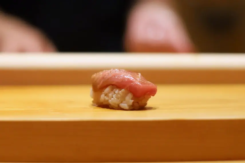 A single piece of tuna nigiri sushi sits on a light wooden counter board. In the softly blurred background, the hands of a chef are subtly visible in the warm lighting.