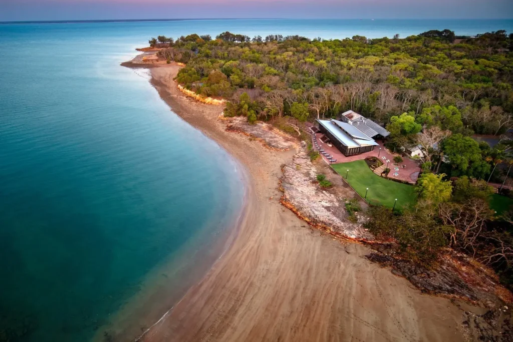 An aerial view of a beachfront resort nestled along a sandy coastline bordered by turquoise ocean waters. A multi-level building with a green lawn sits beside dense green trees and rocky shoreline terrain.