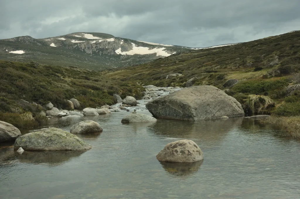 A shallow, stony stream winds through a grassy alpine valley filled with large boulders. In the background, green hills rise toward a distant mountain range with lingering patches of snow under an overcast sky.